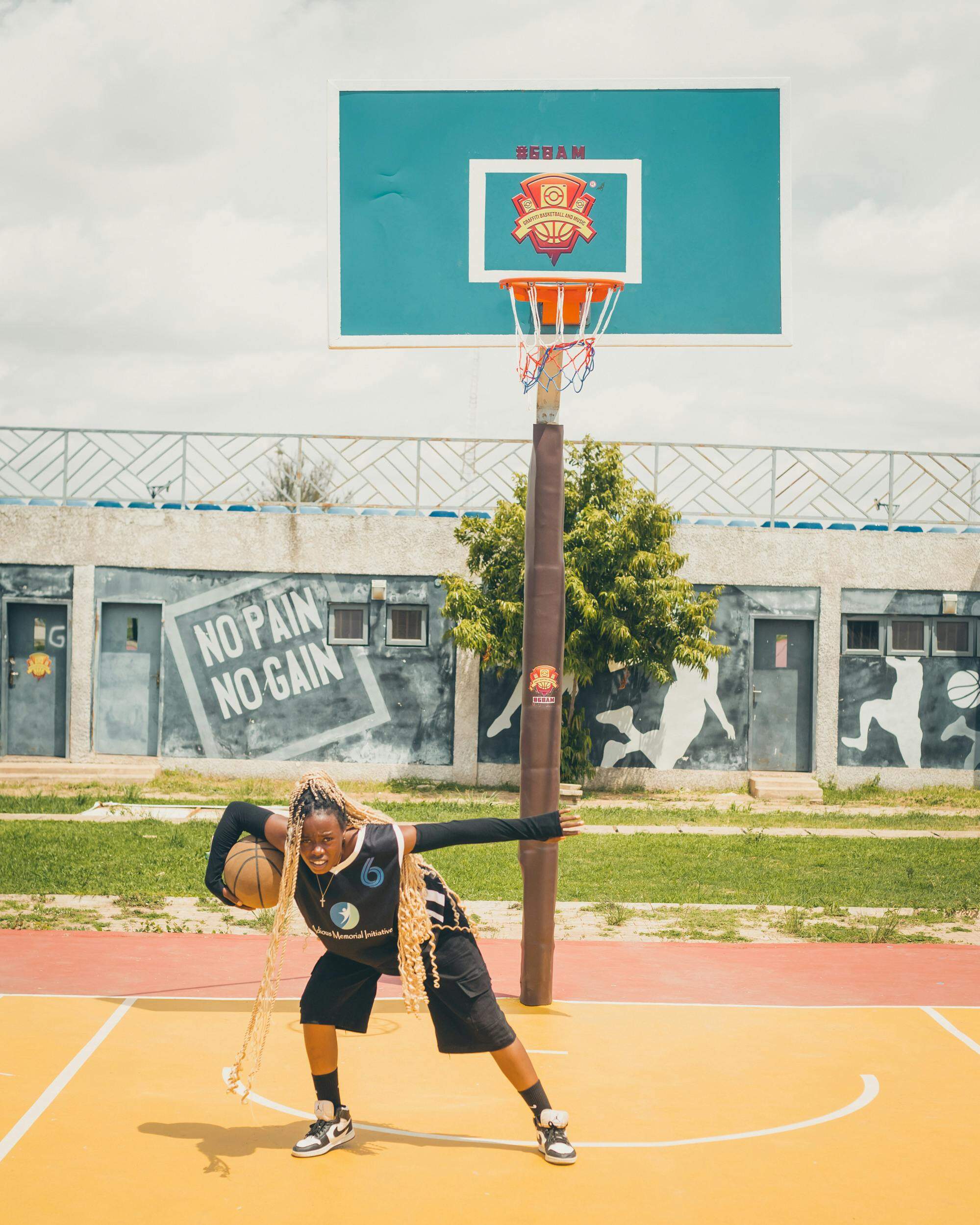 Joueuse de basketball posant avec un ballon sous un panier, sur un terrain extérieur coloré avec une fresque 'No pain no gain