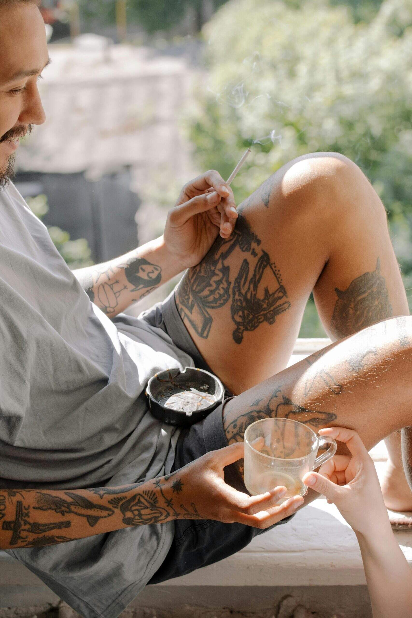 Homme tatoué assis sur un rebord de fenêtre, tenant une cigarette et une tasse, avec un cendrier sur les genoux