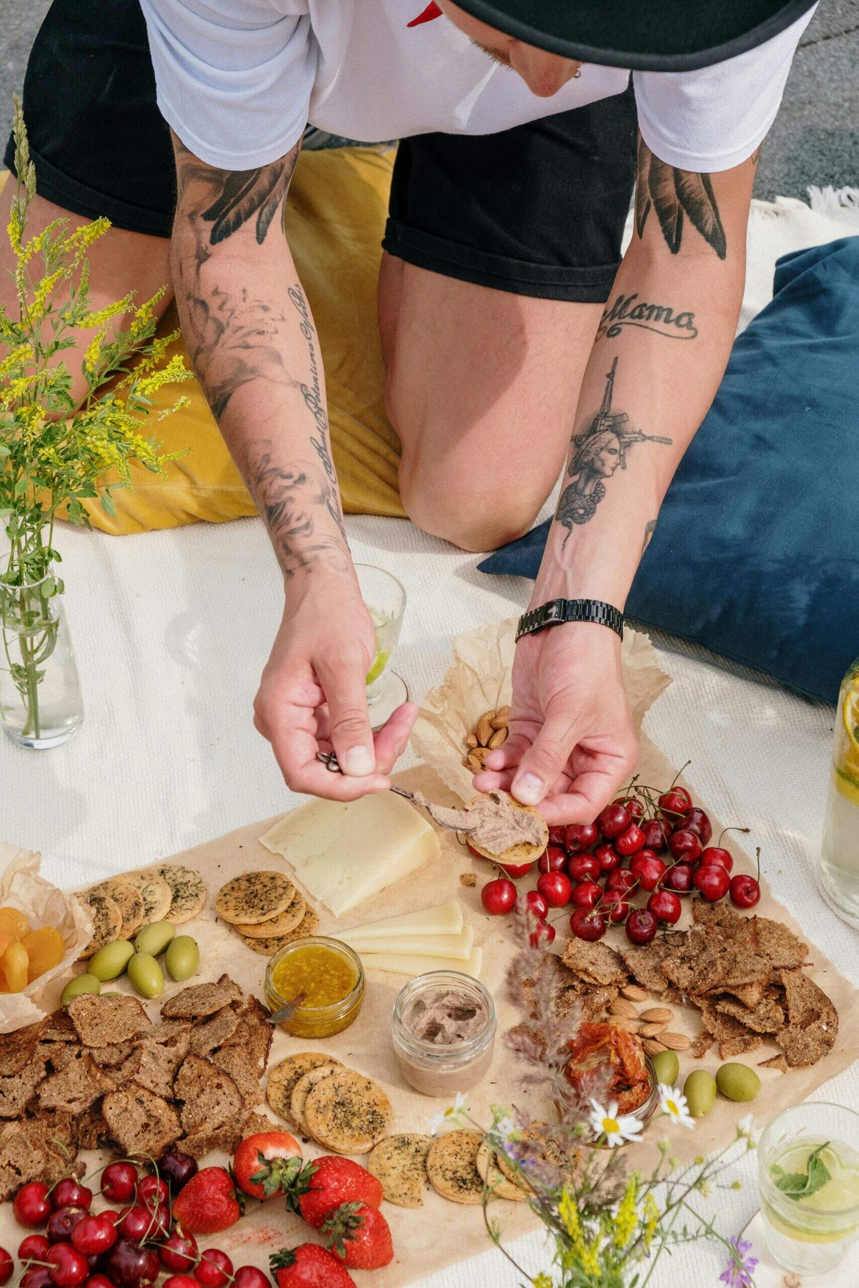Personne tatouée préparant un plateau apéritif avec fruits, fromages et tartinades lors d’un pique-nique