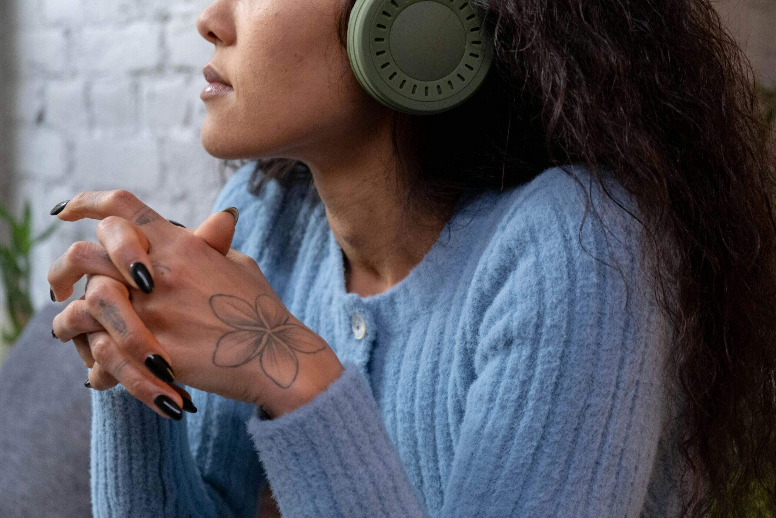 Femme portant un casque audio vert, vêtue d’un pull bleu, les mains jointes avec un tatouage de fleur sur la main.