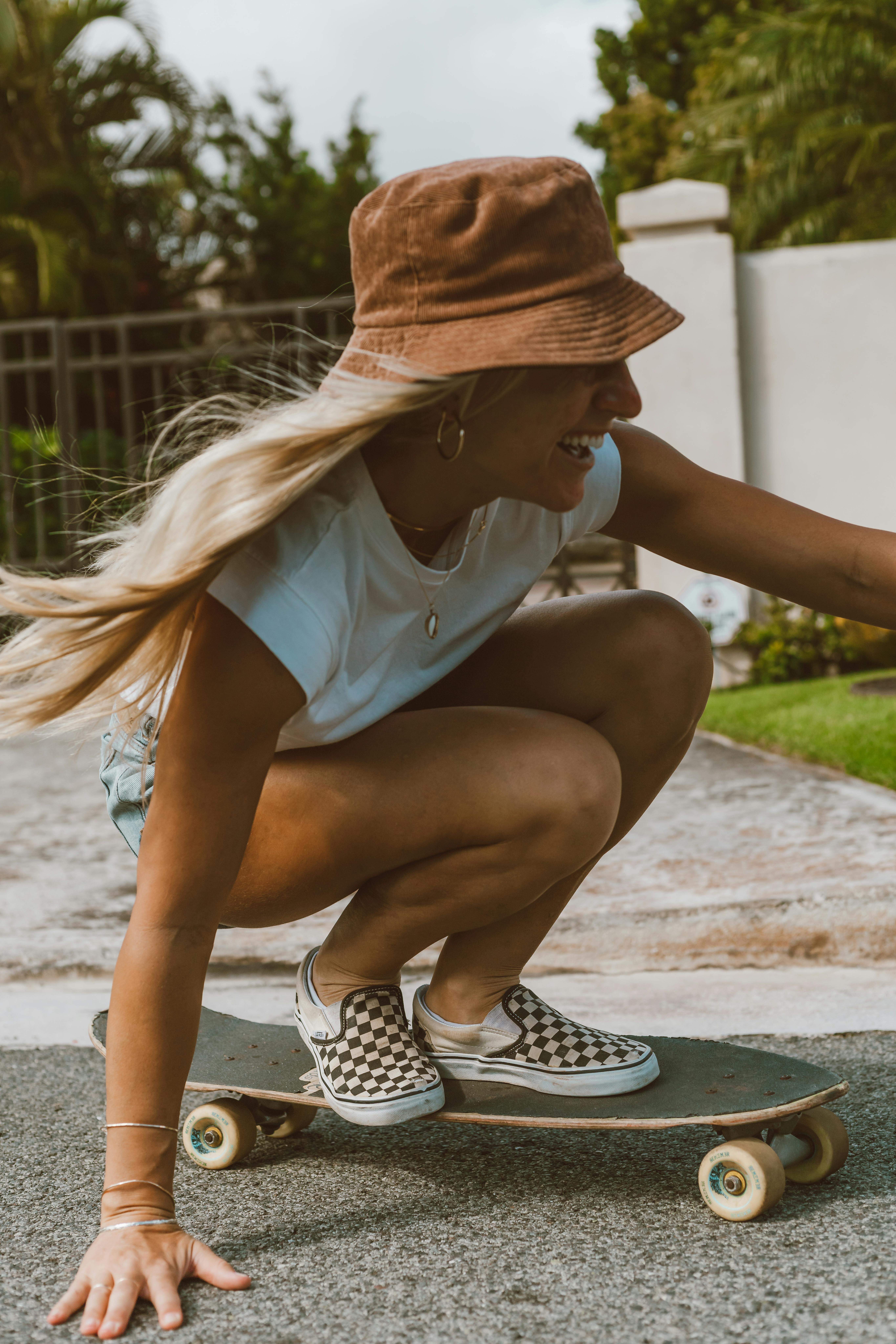 Femme souriante en short et bob brun faisant du skateboard accroupie sur une route résidentielle