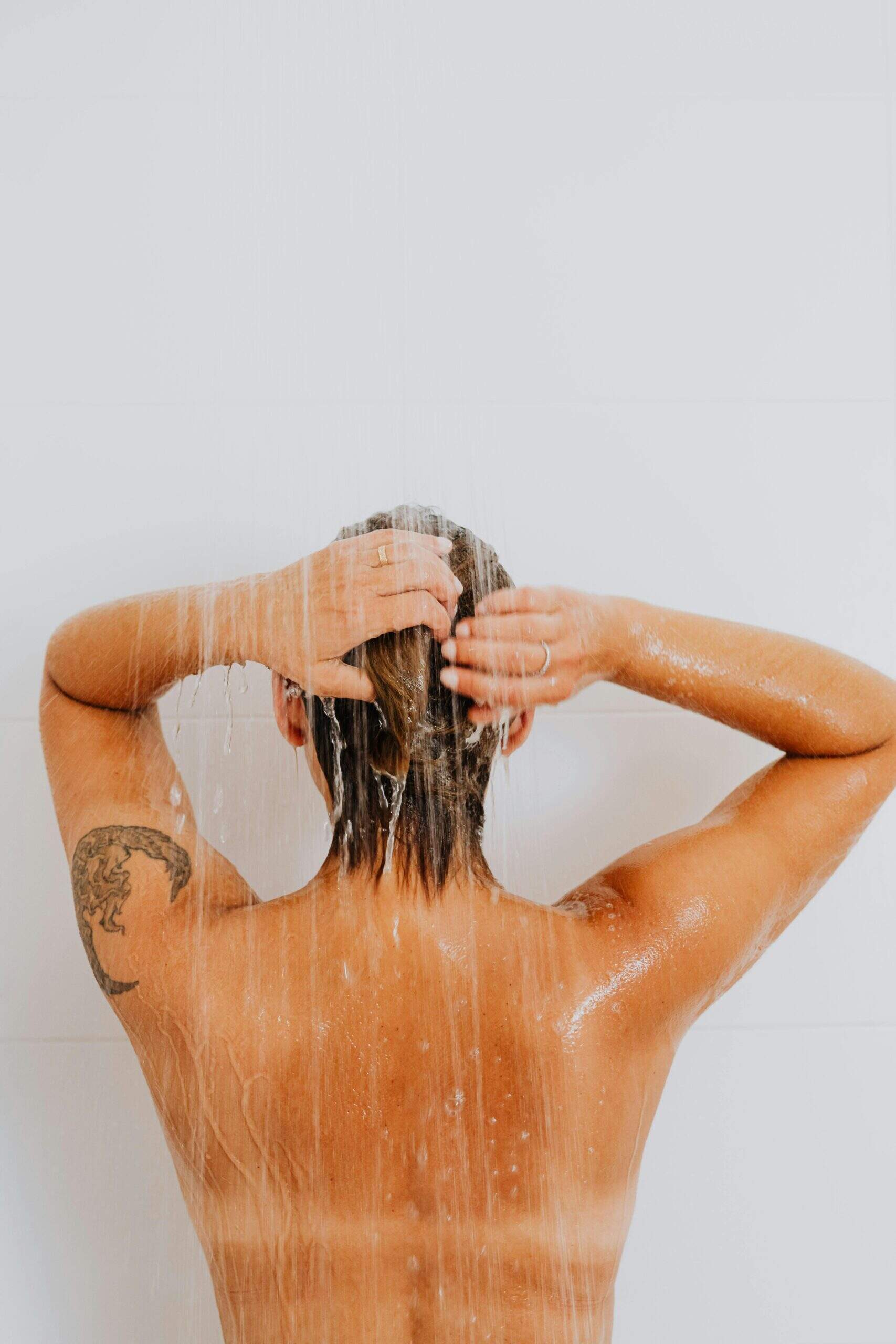 Une personne se lave les cheveux sous la douche, vue de dos, les bras levés et tatouage visible sur le bras.