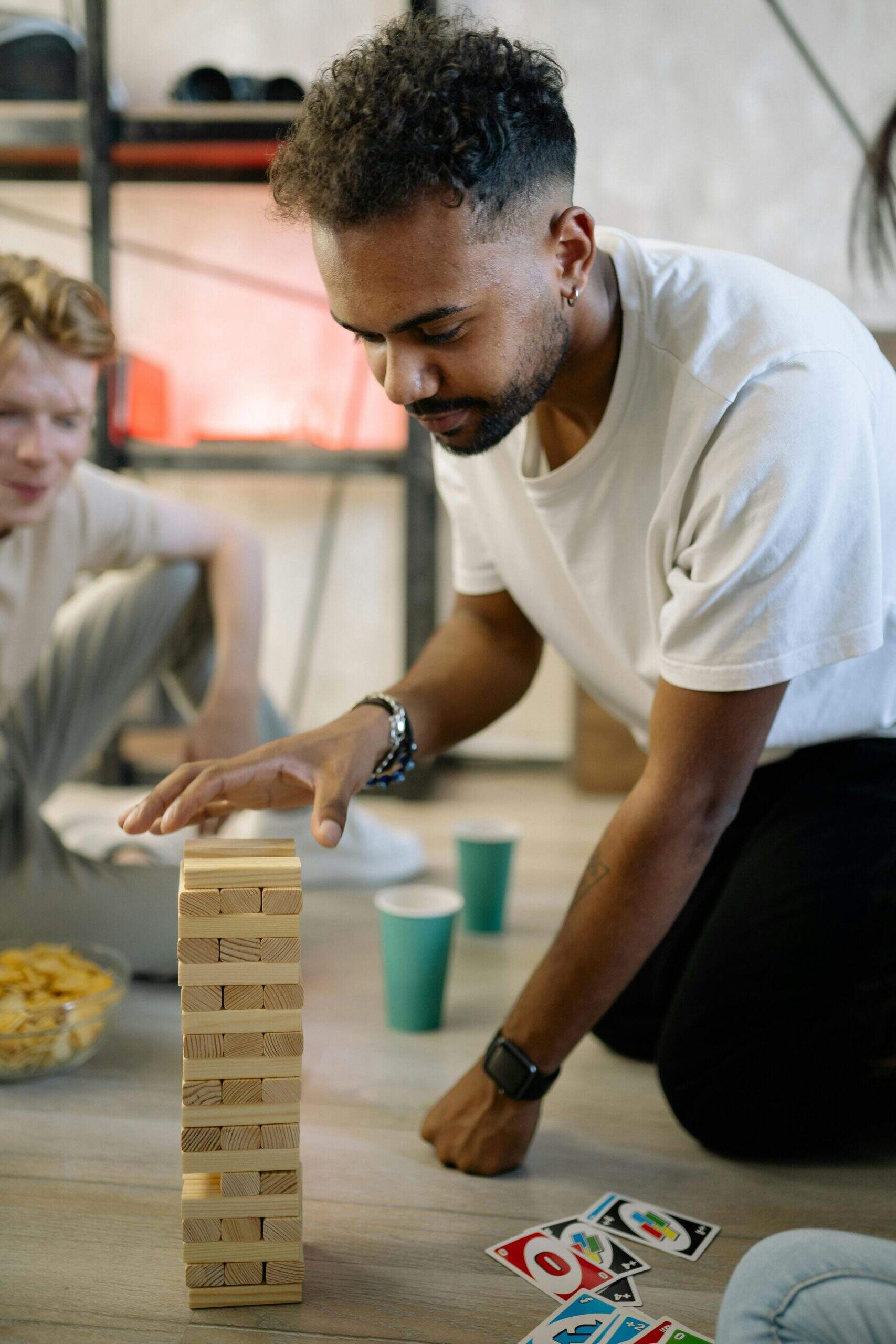 Homme concentré jouant au jeu Jenga, entouré d'amis et de cartes Uno sur le sol