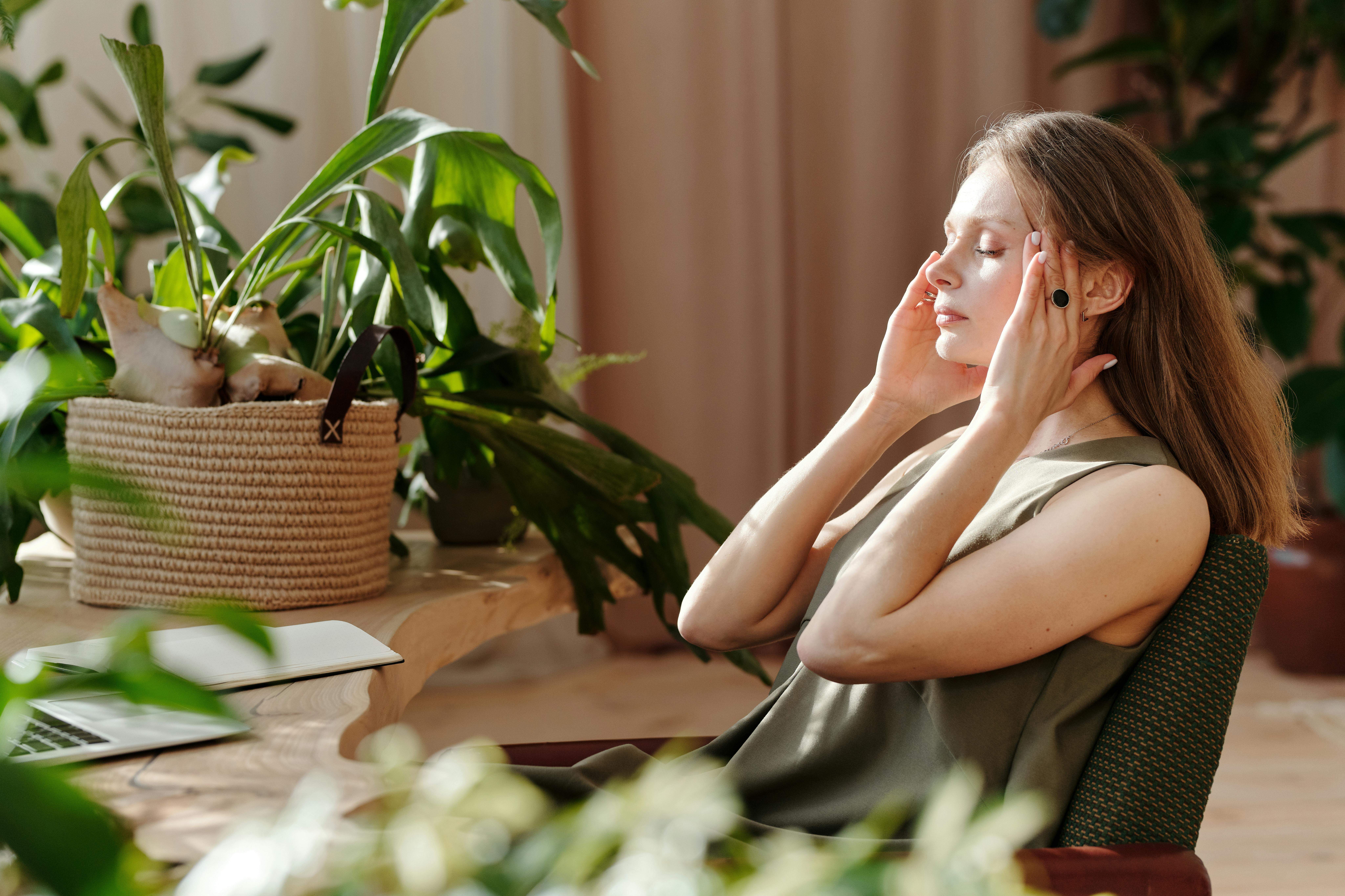 Femme assise dans un intérieur végétalisé, les yeux fermés, les mains sur les tempes.