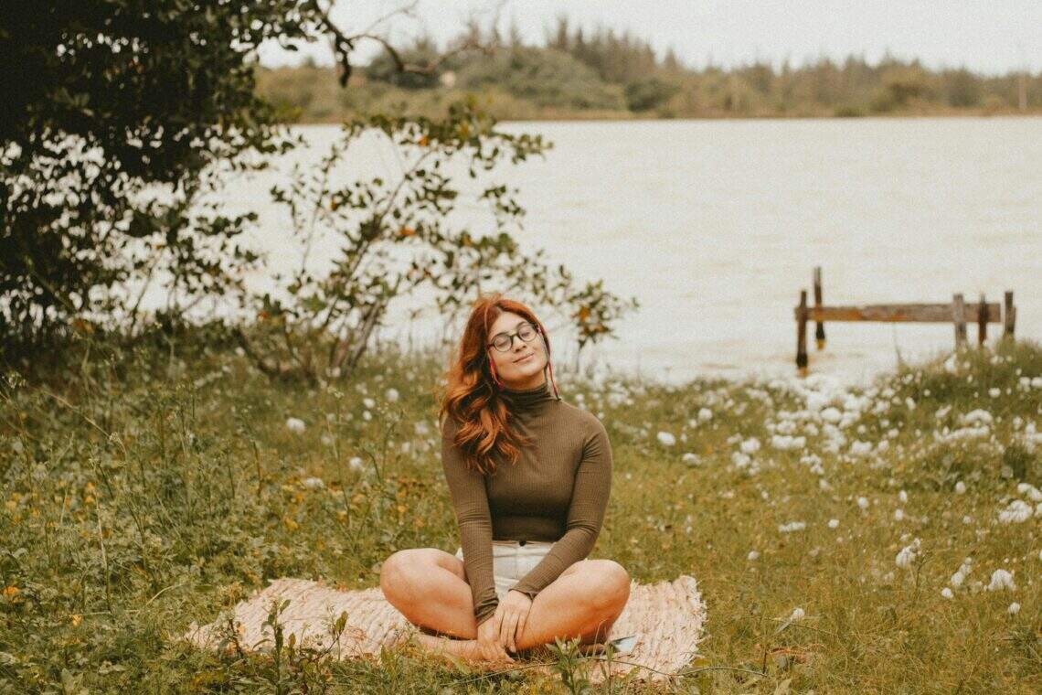 Jeune femme assise sur un tapis en pleine nature, pratiquant la méditation près d’un lac, entourée de fleurs et d’arbres.