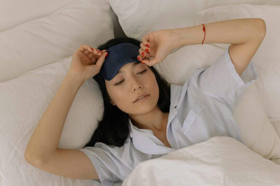 Femme allongée dans un lit blanc, ajustant son masque de sommeil bleu marine